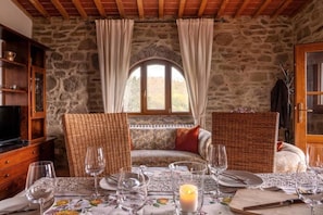 A stone-walled room with a wooden ceiling, a sofa with cushions, a dining table set with glasses and cutlery, and a television on a wooden cabinet.