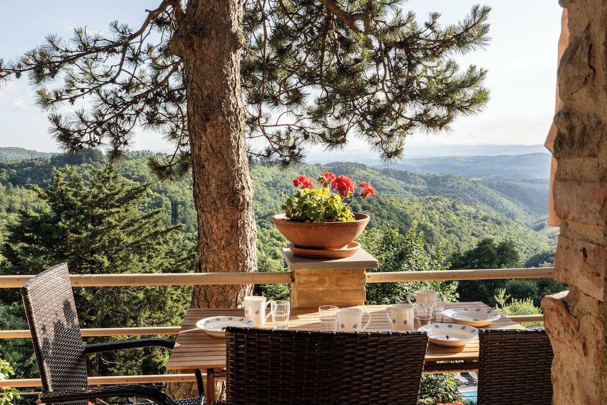 An outdoor dining area with wicker chairs and a table set for two, overlooking a mountainous landscape.