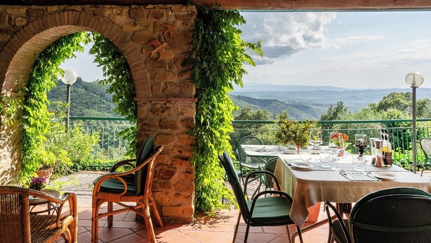 A terrace with stone arches, green plants, and a table set for dining with a scenic mountain view.