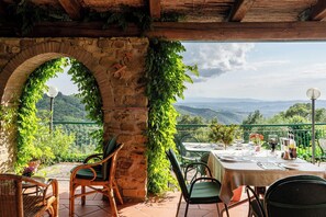 Une terrasse agrémentée d’arcades en pierre, de plantes vertes et d’une table dressée pour les repas, offrant une vue panoramique sur les montagnes.