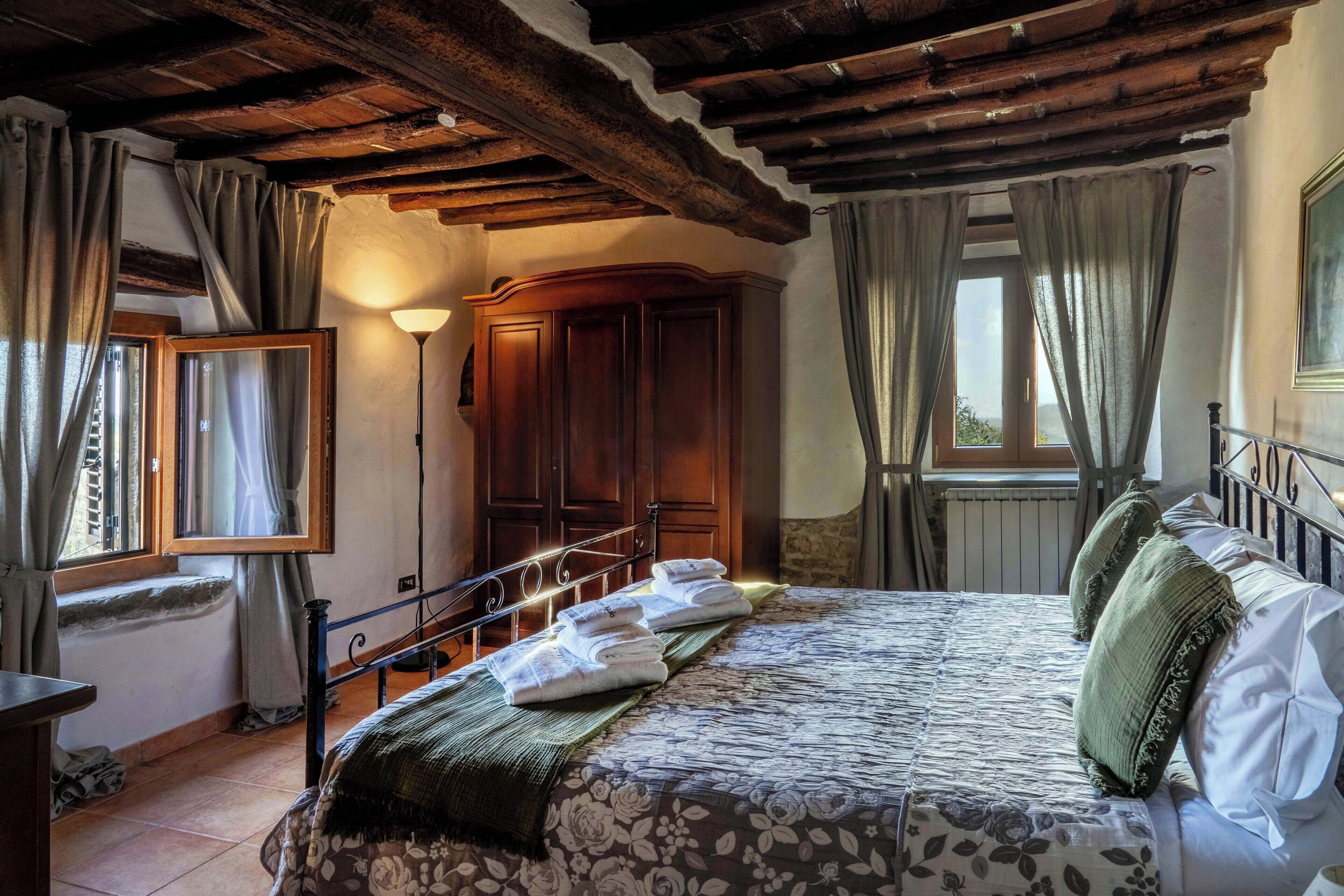 Una camera da letto con soffitto in legno, un letto con copriletto floreale, un comodino, una lampada e una finestra con tende.