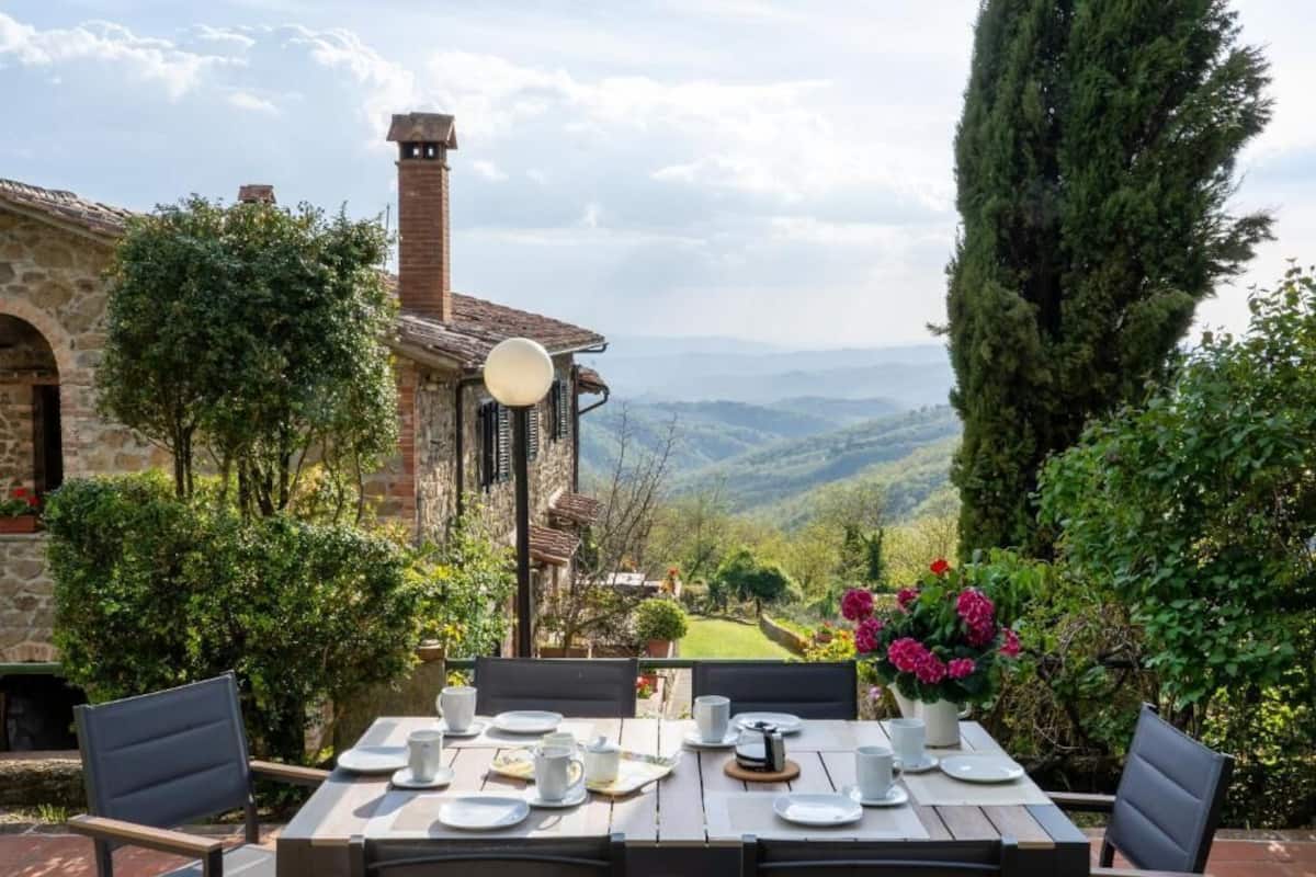 A table set outdoors with a view of a mountainous landscape, a stone building, and a tall tree.