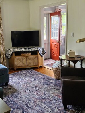 A living room with a flat-screen TV on a wooden cabinet, a patterned rug, a red door, and a small round table with a lamp. - Water Street Retreat (Eastport)