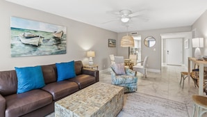 A living room with a brown leather sofa, a patterned ottoman, a painting of boats, a ceiling fan, and a dining area with chairs and a table. - Modern 2BR, 2BA condo in Sarasota's Palm Bay Club, sleeps 6 comfortably. (Sarasota)