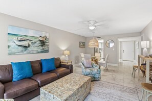 A living room with a brown leather sofa, a patterned ottoman, a painting of boats, a ceiling fan, and a dining area with chairs and a table. - Modern 2BR, 2BA condo in Sarasota's Palm Bay Club, sleeps 6 comfortably. (Sarasota)