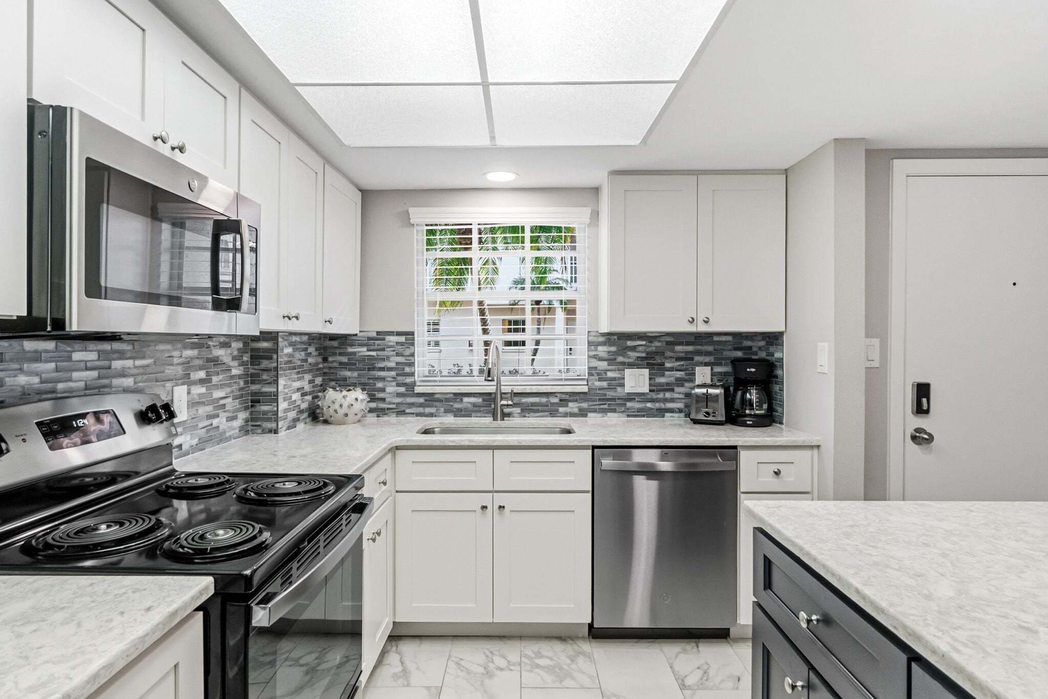 A modern kitchen with a built-in oven, microwave, and dishwasher, a window with a view of greenery, and a tiled backsplash.