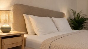 Una cama bien hecha con sábanas blancas, una mesita de noche de madera con una lámpara y una planta en maceta junto a la cama.