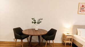 Una habitación con una mesa redonda de madera, dos sillas, una mesita de noche con una lámpara, una cama y un cuadro colgado en la pared.