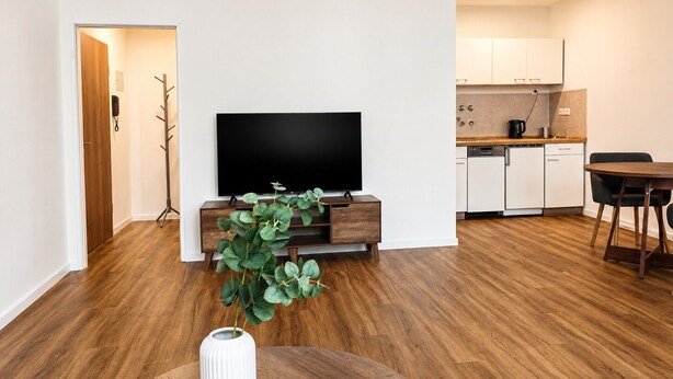 Una sala de estar moderna con televisor de pantalla plana, piso de madera, una zona de comedor con mesa y sillas, y una cocina con armarios blancos.