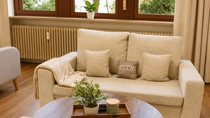 Una sala de estar moderna con un sofá color beige, una mesa de centro de madera y vistas a árboles a través de ventanas grandes.