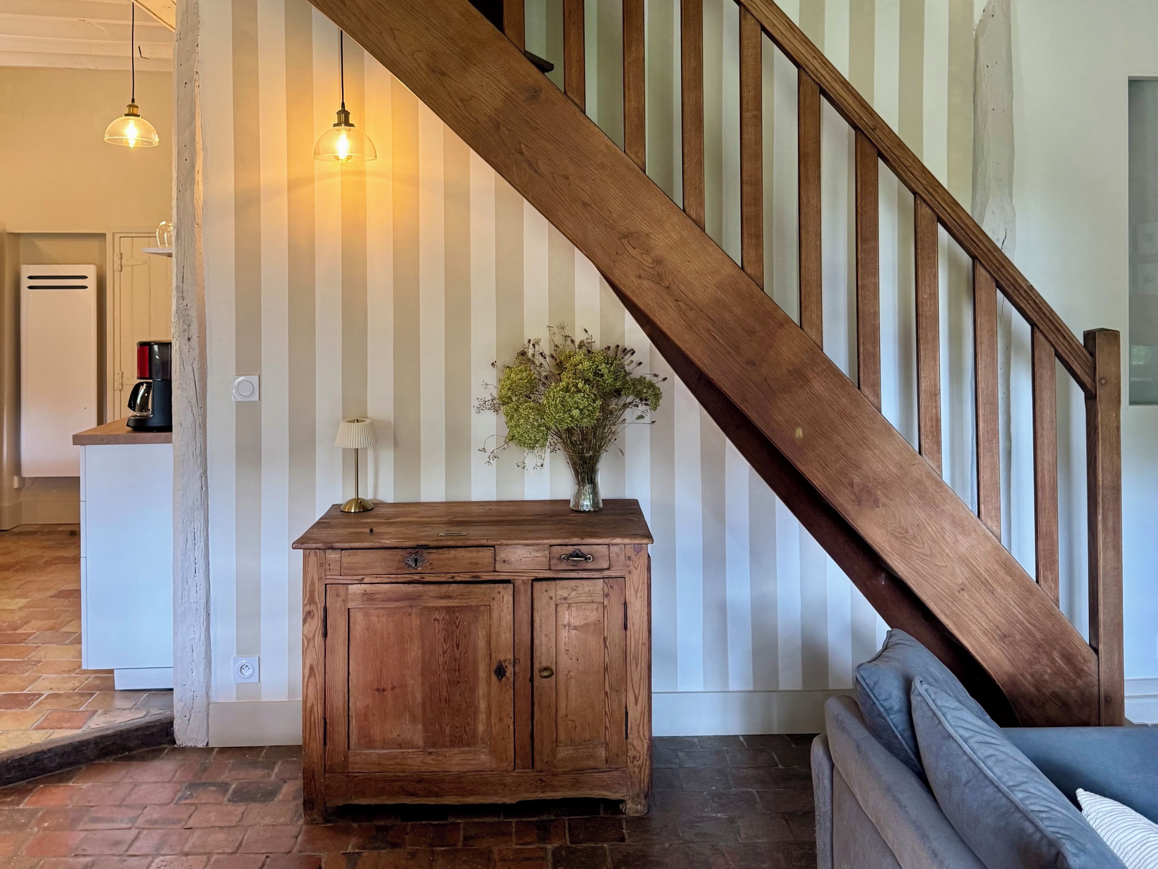 A wooden staircase with a striped wall background, a wooden cabinet with drawers, a lamp, and a vase with flowers.