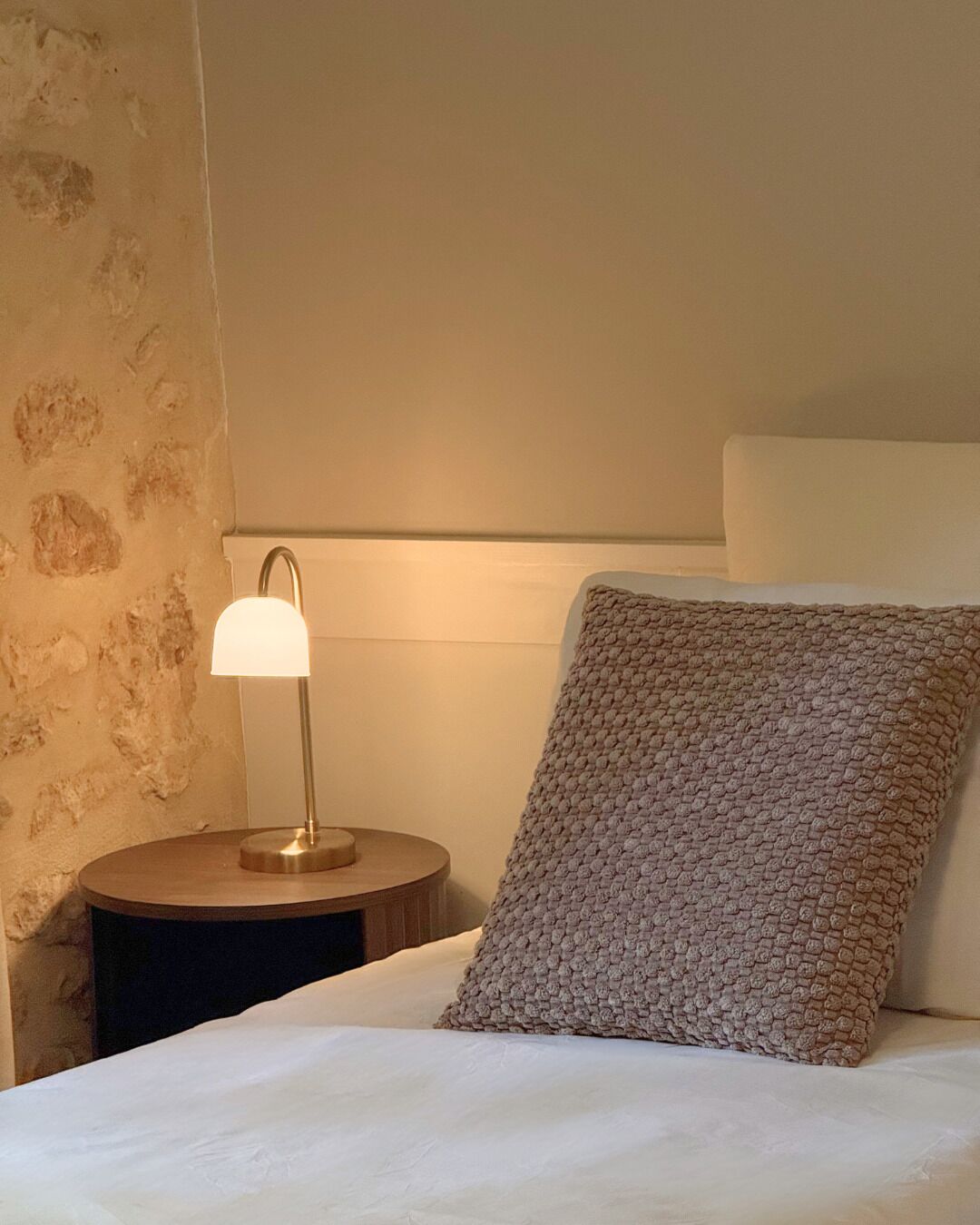Un letto con cuscino bianco e coperta a texture, una lampada da comodino e una parete con motivo effetto pietra.