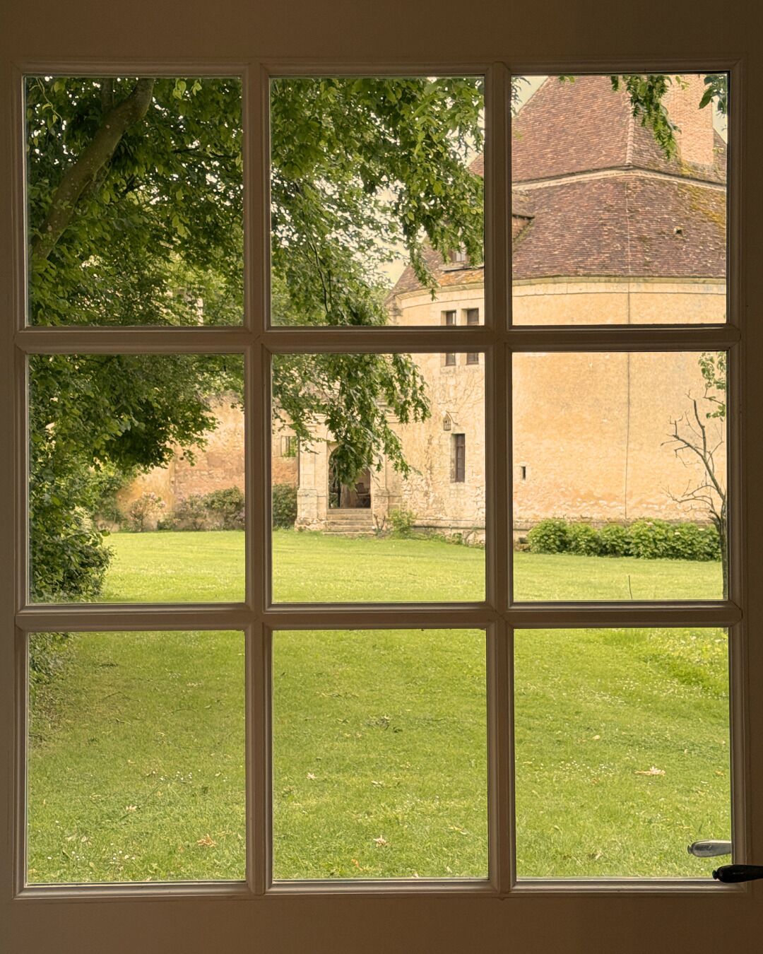 A window with a view of a courtyard, stone building, and greenery.