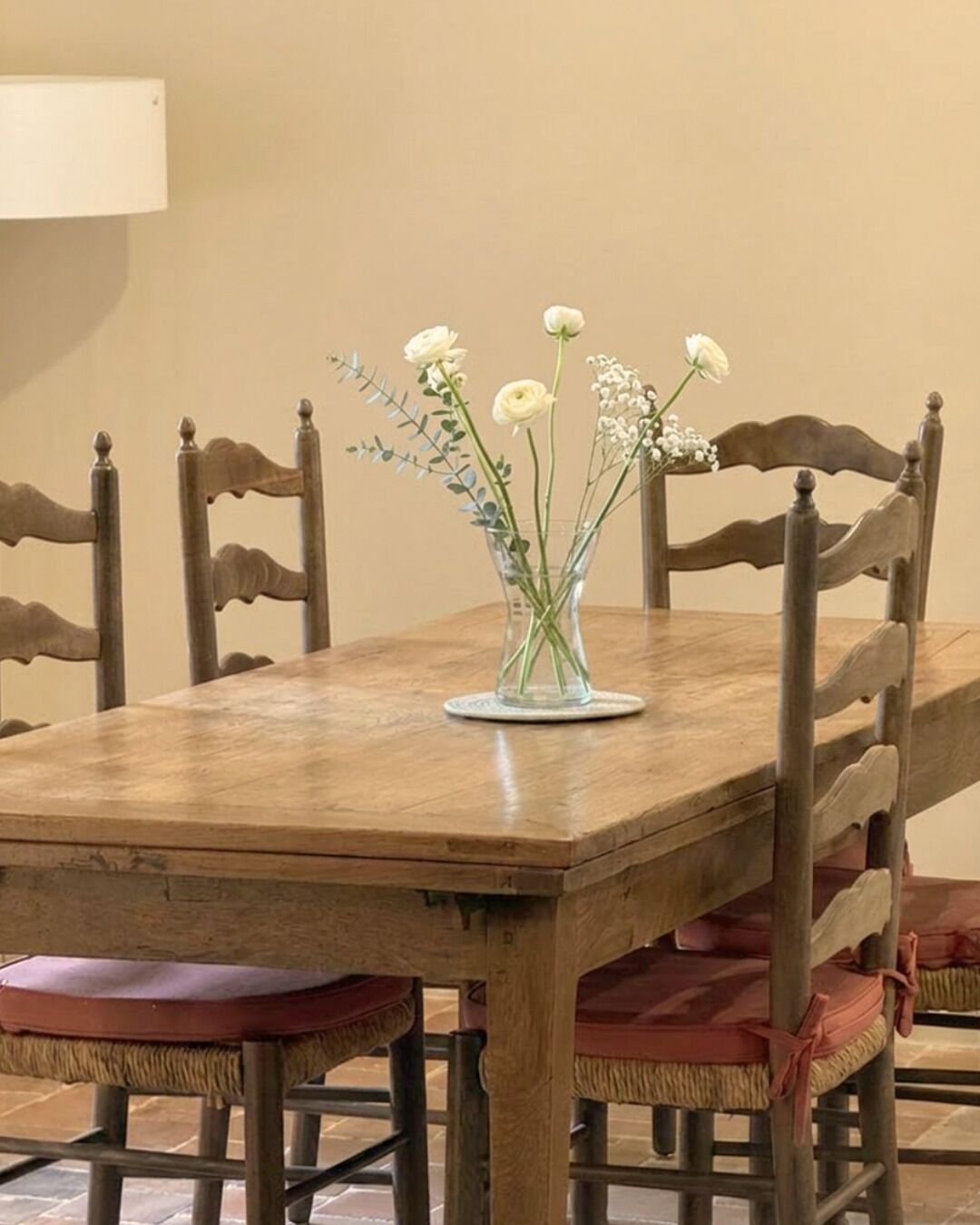 Une table à manger en bois sur laquelle est posé un vase en verre transparent contenant des fleurs blanches et des feuilles vertes.