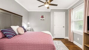 A bedroom with a bed, a ceiling fan, a nightstand, a lamp, a door, and a window with curtains.