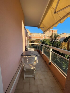 A balcony with a table and chairs, a yellow awning, and a view of apartment buildings. - GOLFE-JUAN (06220) WEEKLY STUDIO RENTAL – JULY & AUGUST (Vallauris)