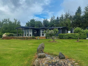 Ein modernes Haus mit überdachter Veranda, Rasen und Garten mit einem kleinen Baum und Steinen.