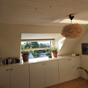 A room with a sloped ceiling, a window with a view of greenery, a potted plant on the windowsill, and a hanging light fixture. - A lush, authentic setting surrounded by nature, yet close to both the city and the archipelago! (saltsjo-boo)