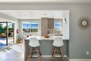 A modern kitchen with a marble countertop, two bar stools, and a view of the outdoors. - Salt & Sun Palm Springs by Navigate Stays (Palm Springs)
