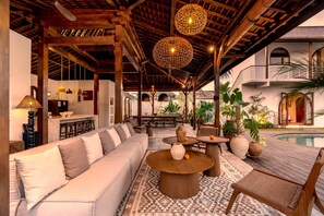 Un área de estar exterior techada con piscina, que incluye un sofá, mesas redondas e iluminación esférica colgante.