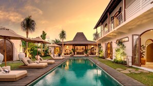 A pool area with lounge chairs and umbrellas, a dining area with a thatched roof, and a modern building with large windows. - Villa Amara in Kuta with 7 bedrooms and 7 bathrooms (Kuta)