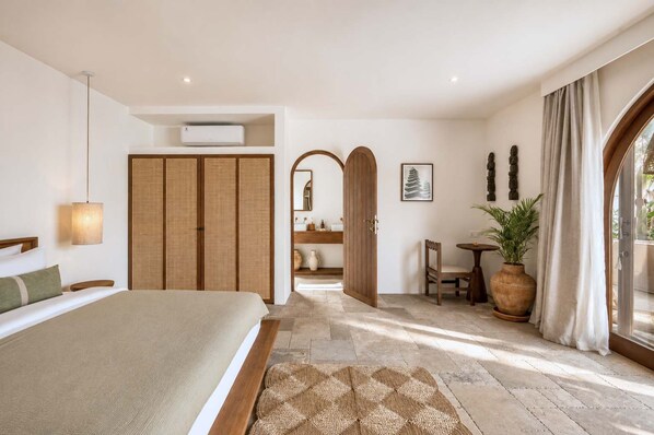 Ein Schlafzimmer mit Bett, Nachttisch, Spiegel, Kleiderschrank, einem Badezimmer mit Waschbecken und Spiegel, einem kleinen Tisch mit Pflanze und einem großen Fenster mit Vorhängen.