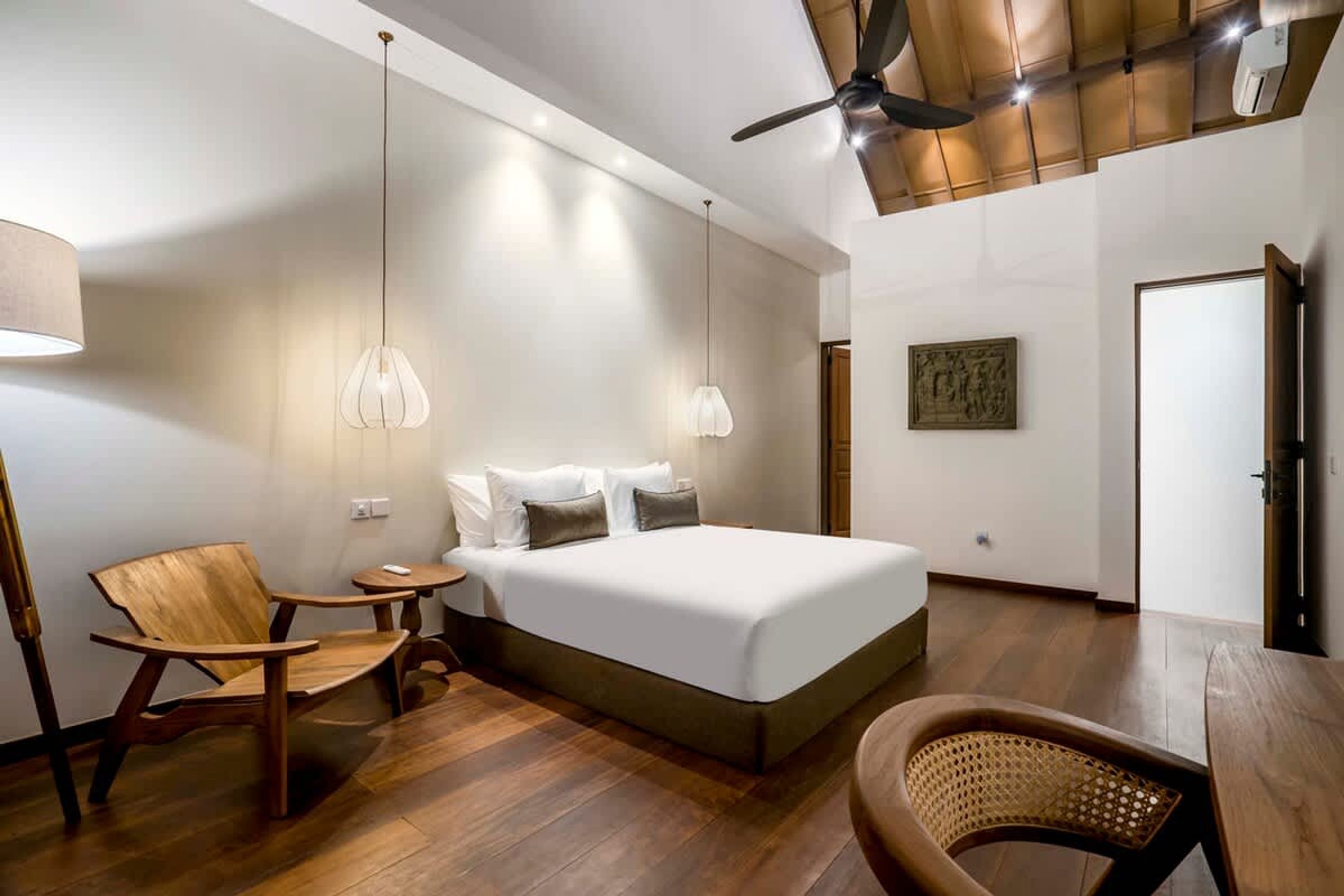 A bedroom with a bed, wooden chairs, a table, a ceiling fan, and a wall-mounted air conditioner.