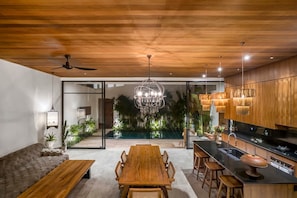 A modern kitchen with wooden cabinets, a dining area with a wooden table and chairs, and a view of a pool. - Nusantara Loft Alor in Pererenan (Pererenan)