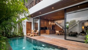 Et moderne hus med swimmingpool, træterrasse og et køkkenområde, der kan ses igennem glasdøre.