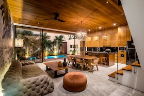 A modern kitchen with wooden cabinets, a dining area with a table and chairs, a sofa, and a staircase. - Nusantara Loft Raijua (Pererenan)