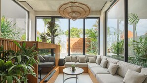 Ein modernes Wohnzimmer mit Sofa, Couchtisch und Fernseher. Große Fenster und Pflanzen sind vorhanden.