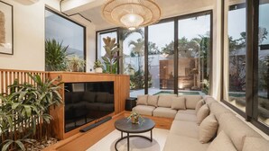 Ein modernes Wohnzimmer mit einer Eckcouch, einem runden Couchtisch und einem Fernseher. Große Fenster, eine Wand aus Holzpaneelen und verschiedene Pflanzen sind vorhanden.