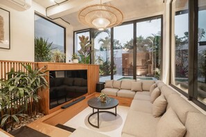 Ein modernes Wohnzimmer mit einer Eckcouch, einem runden Couchtisch und einem Fernseher. Große Fenster, eine Wand aus Holzpaneelen und verschiedene Pflanzen sind vorhanden.