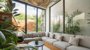 Ein modernes Wohnzimmer mit einem großen Ecksofa, einem runden Couchtisch und großen Fenstern mit Blick auf Grünflächen und eine Backsteinmauer.