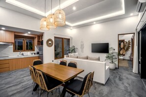 Eine moderne Küche mit Holzausstattung, einem Essbereich mit Holztisch und Korbstühlen, einem an der Wand montierten Flachbildfernseher und einem Blumentopf.