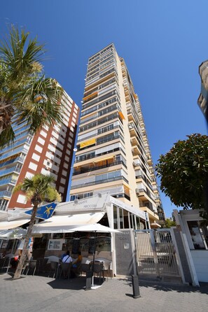 Ein hohes Gebäude mit mehreren Balkonen, einem Restaurant mit Terrassenplätzen und einer Palme.