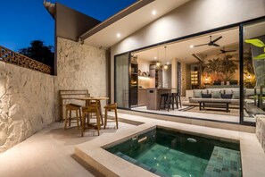 A modern outdoor living area with a pool, seating, and a kitchen visible through glass doors. - Sunrise Lush River Villa (Pererenan)