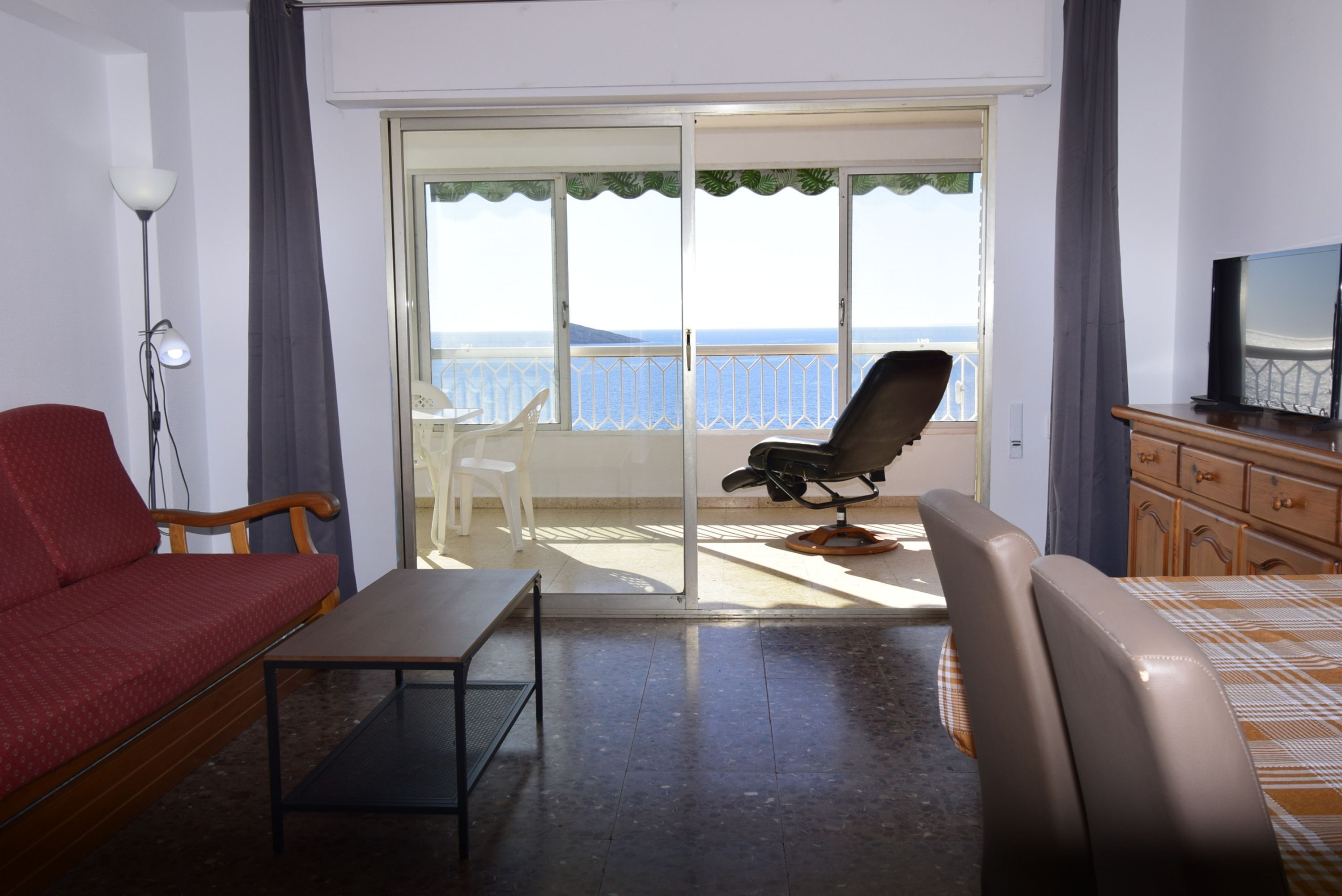 A room with a sofa, a chair, a small table, a TV, and a balcony with a view of the sea.