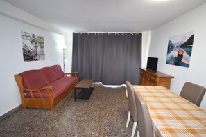 A room with a sofa, a small table, a TV stand, and a bed with a checkered bedspread. - Torre Principado 20E (Benidorm)