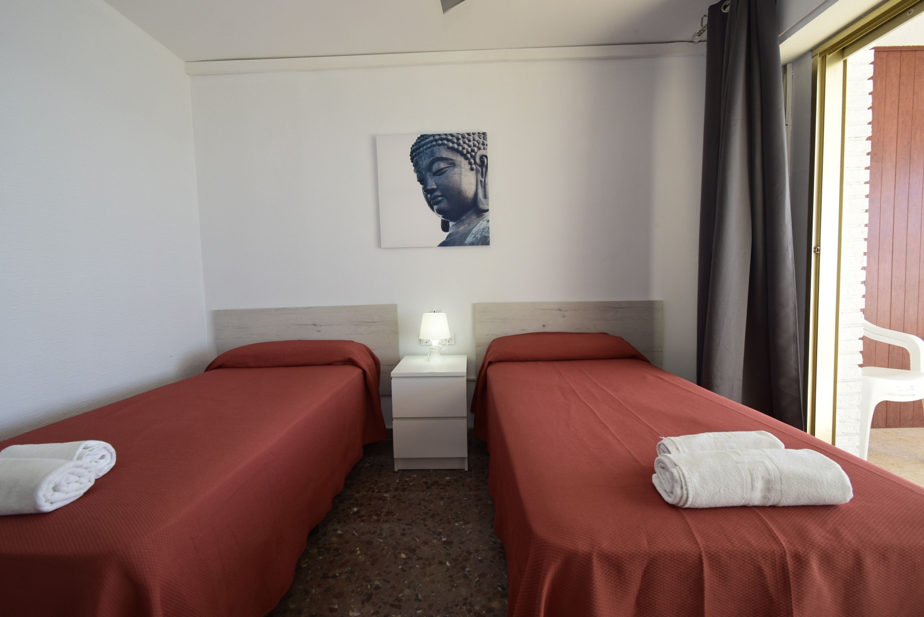 A hotel room with two single beds, a nightstand with a lamp, a Buddha painting on the wall, and a view of the outside.