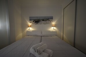 A neatly made bed with white linens and two rolled towels, a wall-mounted artwork depicting a beach scene, and two wall-mounted lamps on either side of the bed. - Allonaya D 2-6 in Benidorm (Benidorm)