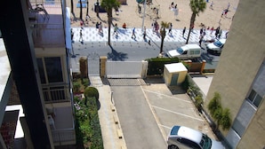 Un estacionamiento con un coche, una entrada con puerta y vista a una playa con gente y palmeras.