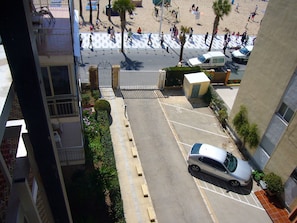Ein Parkplatz mit einem Auto, einem verschlossenen Eingang und Blick auf einen Strand mit Personen und Palmen.