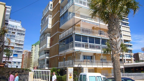 Un edificio residencial de varios pisos con balcones, una entrada protegida por una reja y una palmera en primer plano.