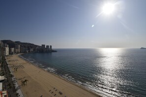 Ein Strand mit blauem Himmel und Sonne, ein sandiger Uferstreifen und Gebäude entlang der Küste.
