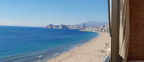 A beach view from a balcony with a clear sky, a sandy beach, and a cityscape in the distance.