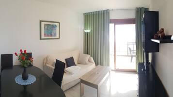 Una habitación con una mesa de comedor, un sofá, un televisor en un mueble bajo y una ventana con cortinas.