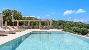 A swimming pool with lounge chairs and a pergola, surrounded by greenery and a clear sky. - Thaloria Villa (Rethymno)
