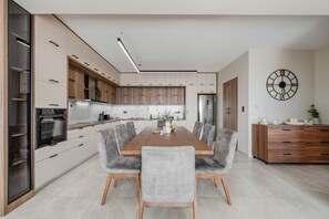 A modern kitchen with a central island, built-in appliances, and a dining area with grey upholstered chairs. - Thaloria Villa (Rethymno)