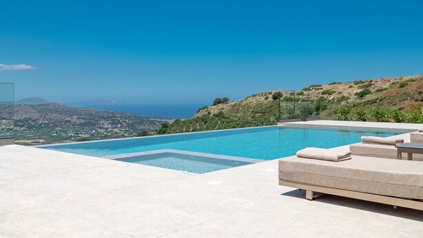 A swimming pool with a view of the sea and hills, a lounge area with cushions, and a clear sky. - Thaloria Villa (Rethymno)
